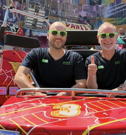 Zwei im Praxis-Shirt mit grünen Sonnenbrillen in einem Fahrgeschäft auf dem Johannisfest